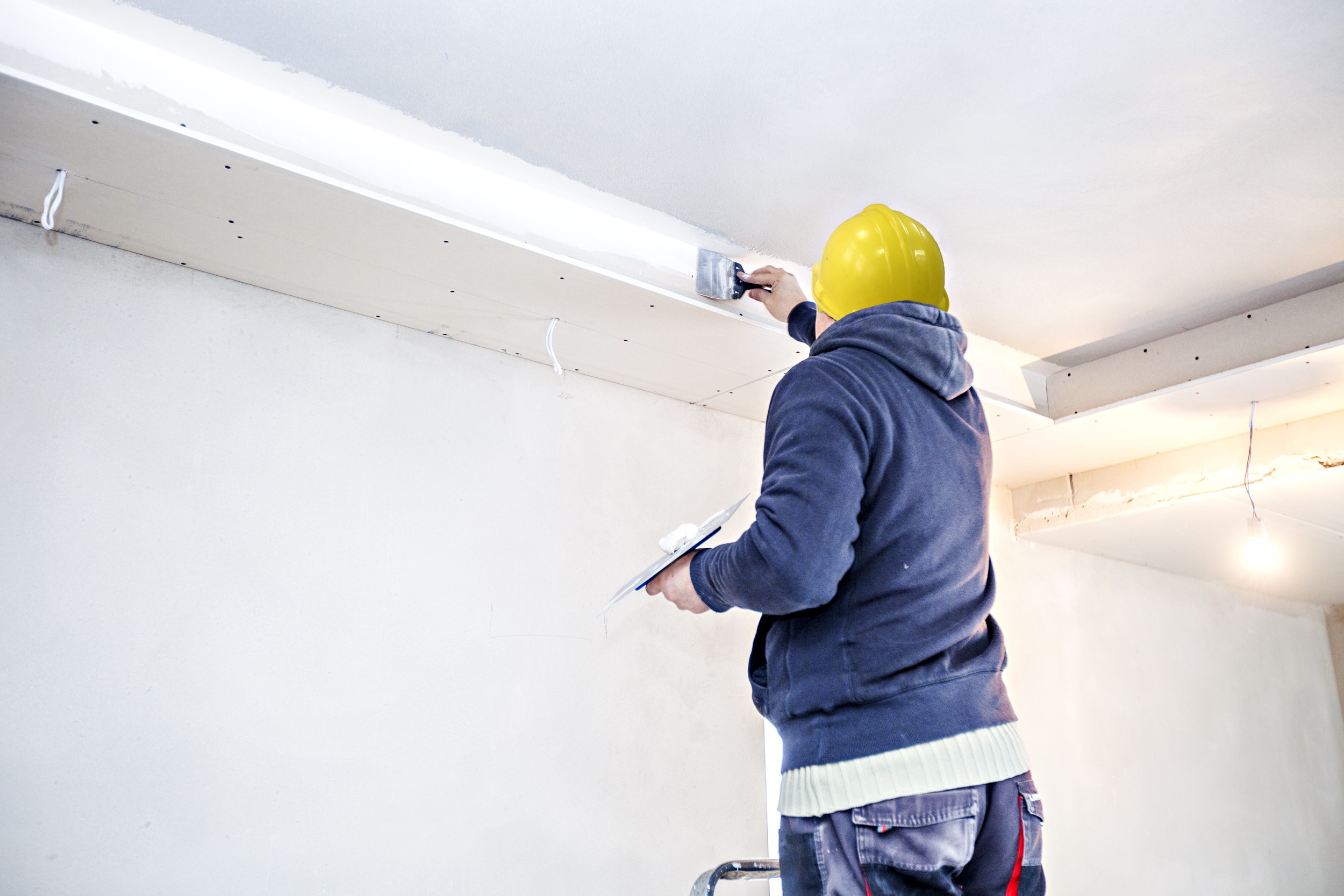 A construction worker with a yellow hard hat paints the ceiling of an unfinished room, holding a paint roller and a trowel.