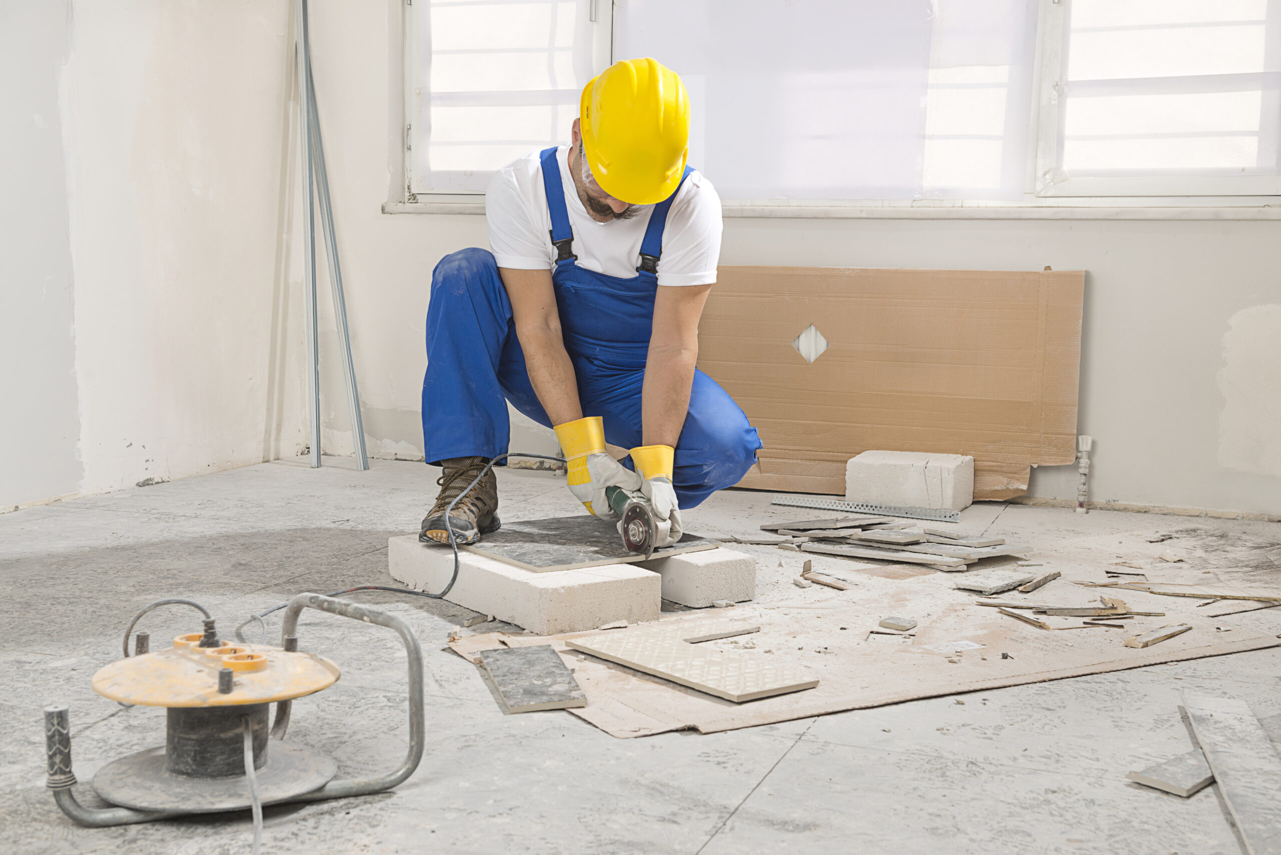 A construction worker in blue overalls and a yellow hard hat uses a power tool on a concrete block in a renovated room with scattered debris.