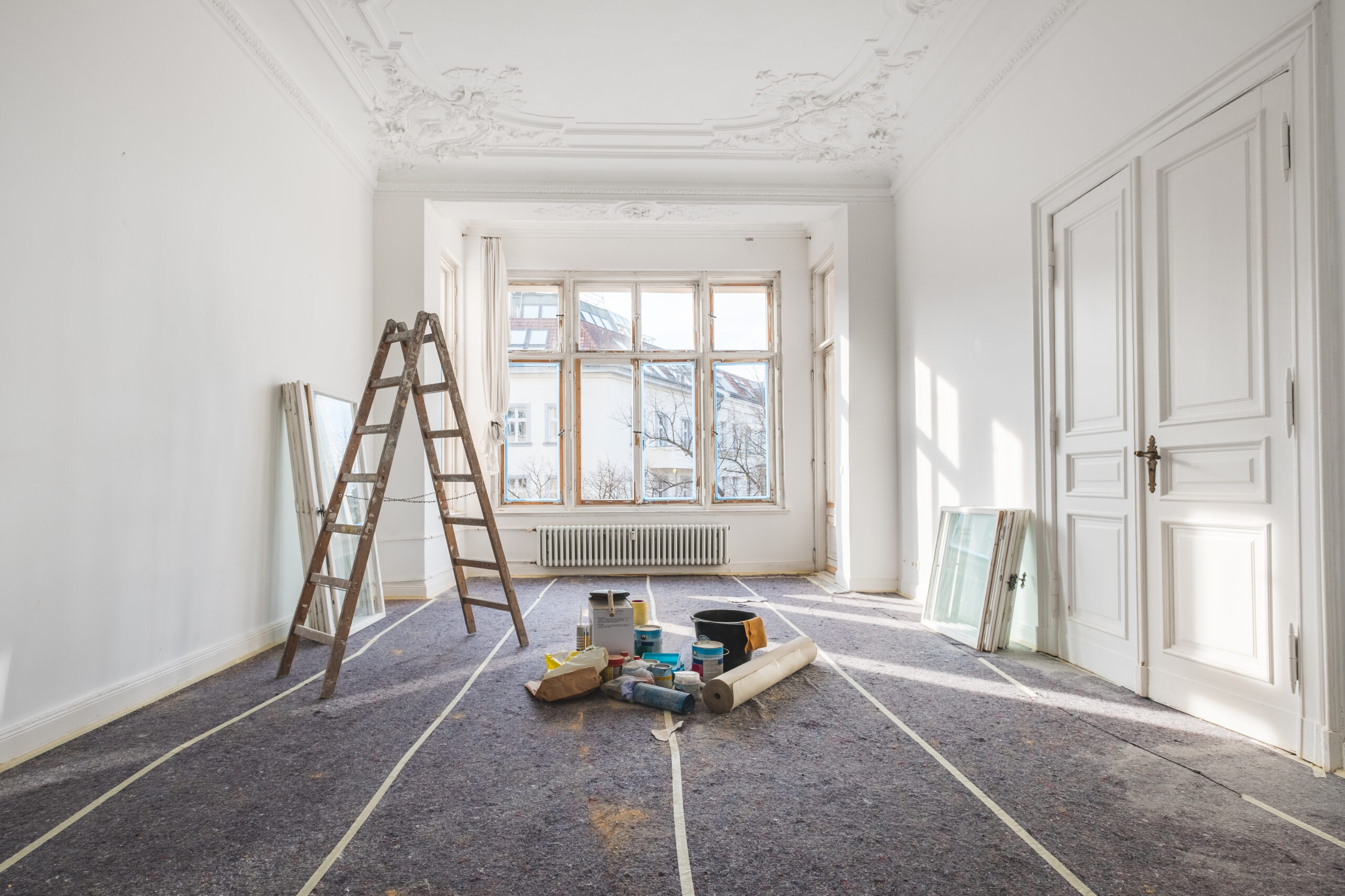 A bright, empty room with a wooden ladder and various painting supplies on the floor. Sunlight streams through large windows, highlighting renovation work.